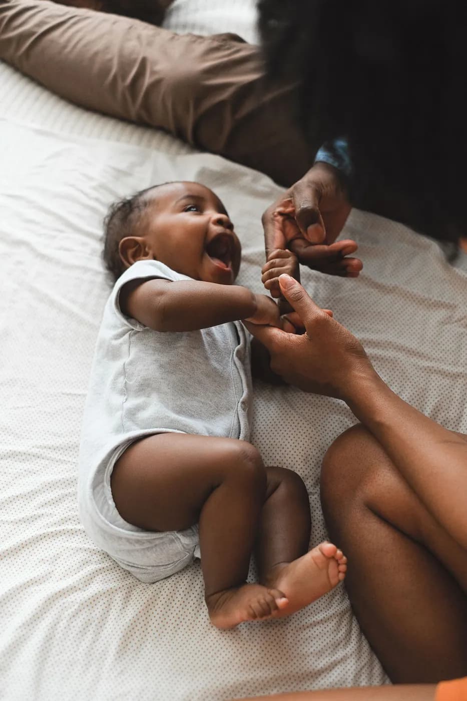 A parent holding a laughing baby's hands on a bed
